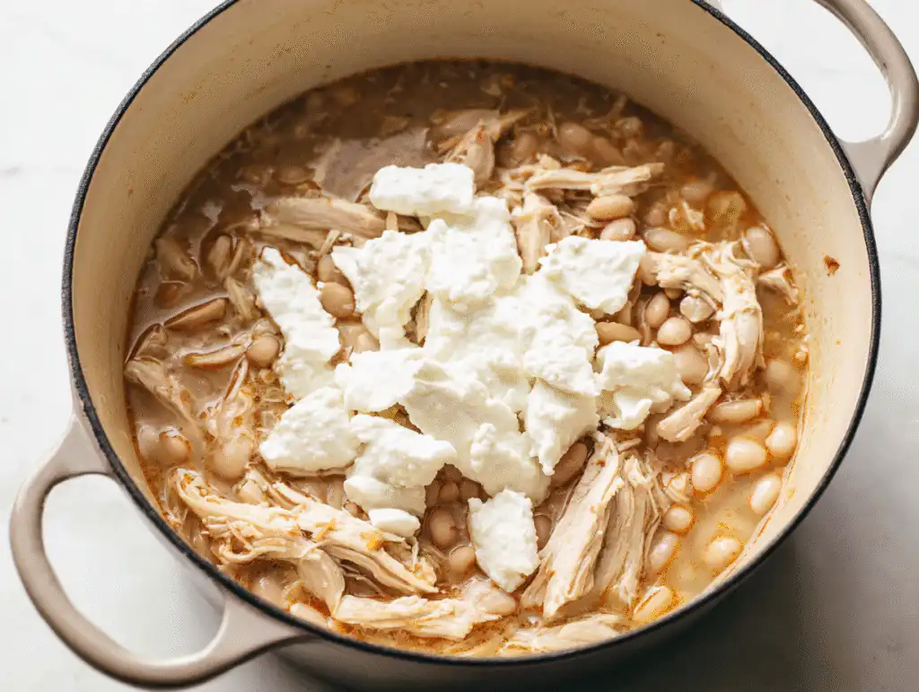 Adding cream cheese to the creamy white chicken chili in the pot.